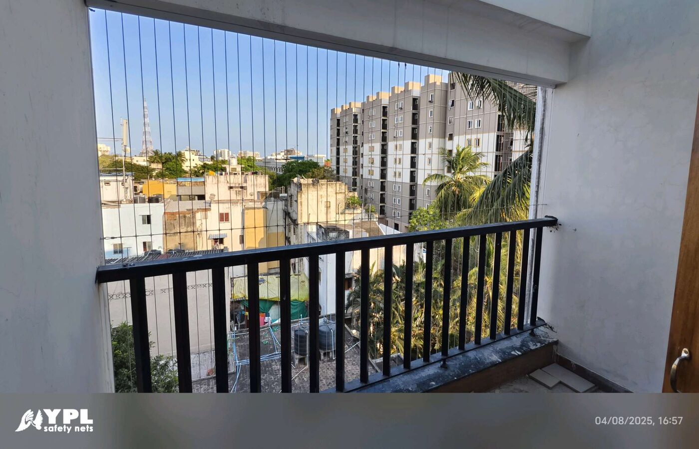 White-walled balcony with invisible grill wires and black railing overlooking coconut palms and nearby apartment buildings.