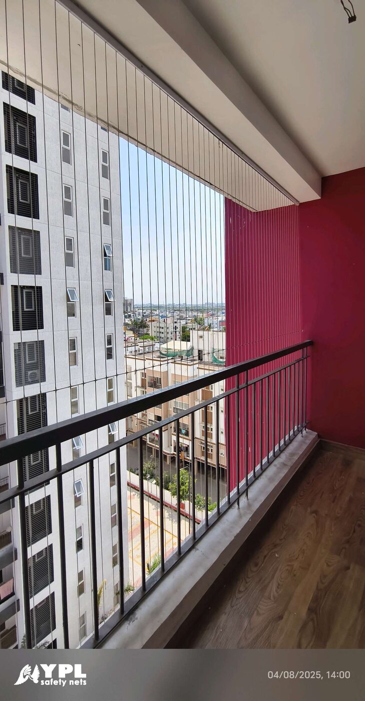 Long balcony with invisible grill wires, black railing, wood-look flooring, and red wall beside a neighboring high-rise.