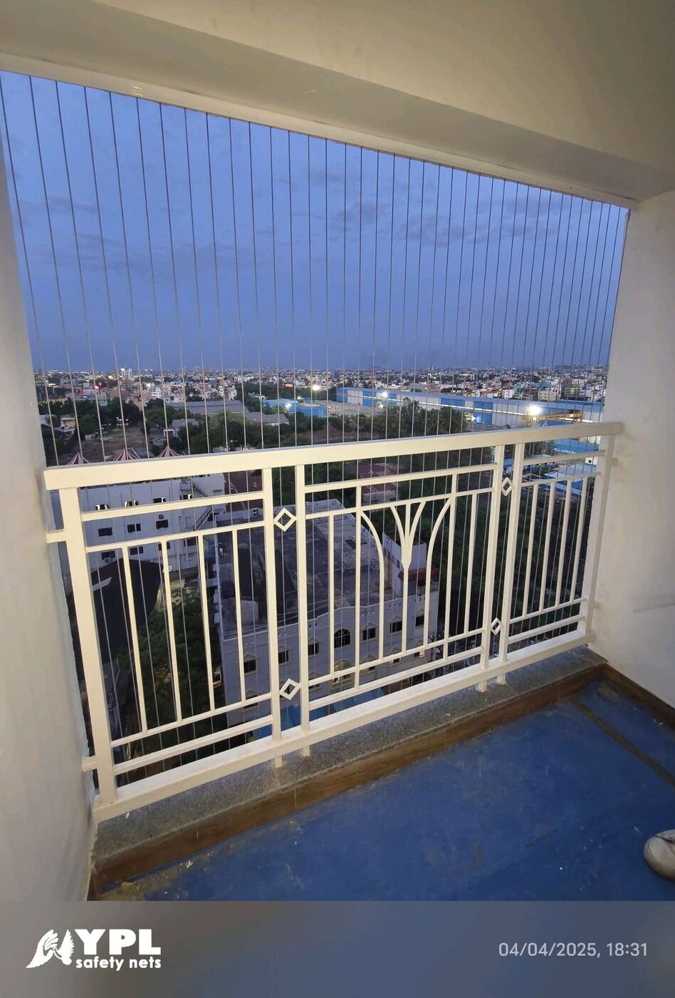 Balcony with vertical invisible grills, decorative white railing, and blue floor overlooking city buildings at dusk.