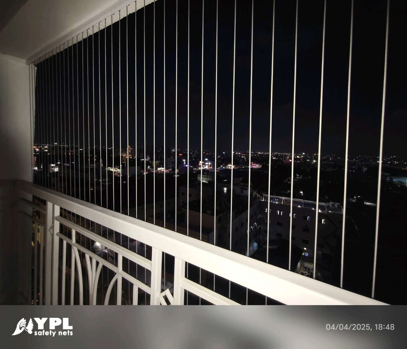 Vertical invisible grills above a white balcony railing, overlooking illuminated city buildings at night.