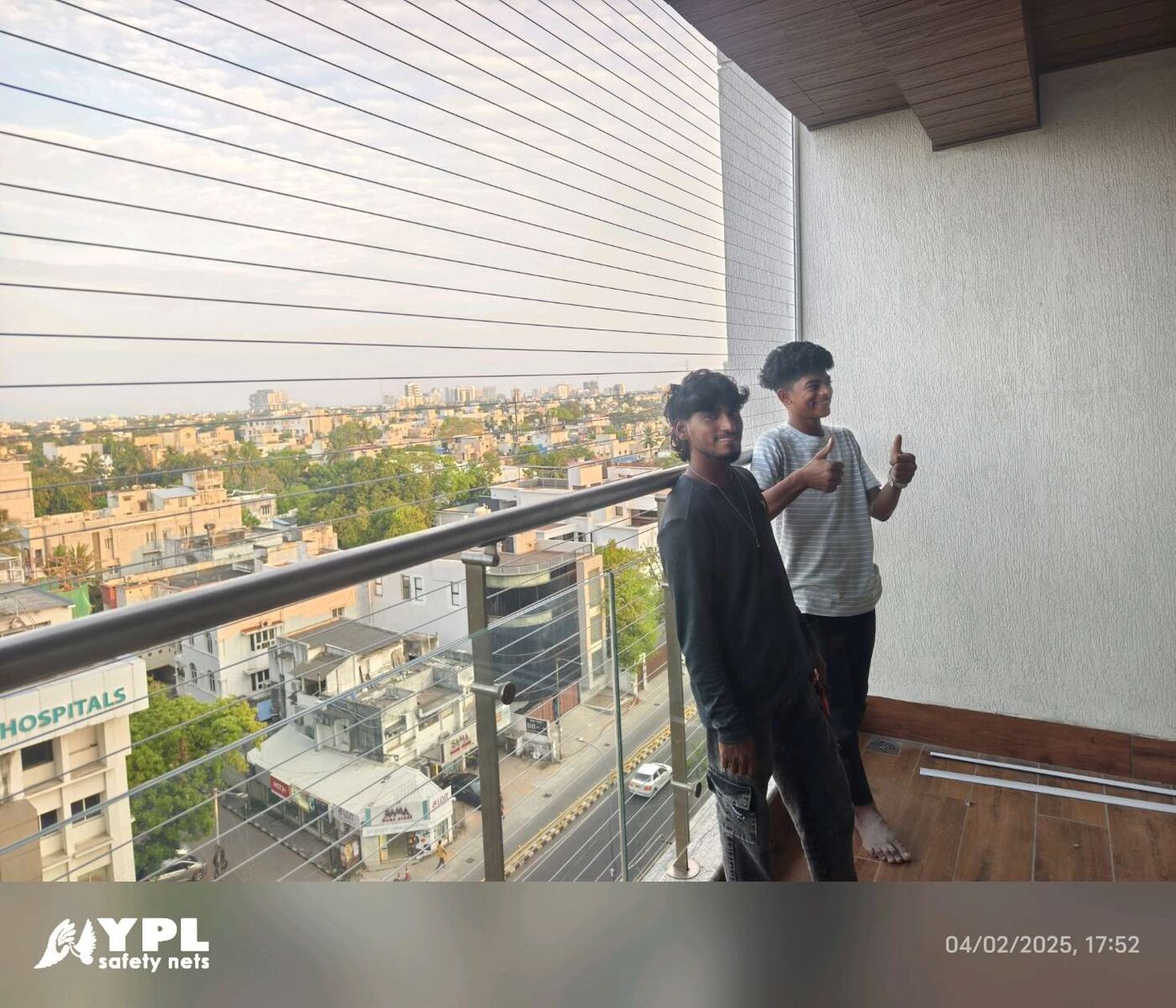 Two people on a wood-floored balcony with horizontal invisible grills and glass railing overlooking a city street.
