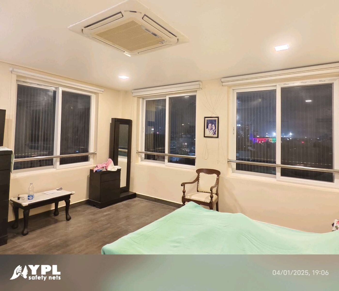Bedroom with three sliding windows fitted with vertical invisible grills, city lights outside, and a ceiling cassette air conditioner.