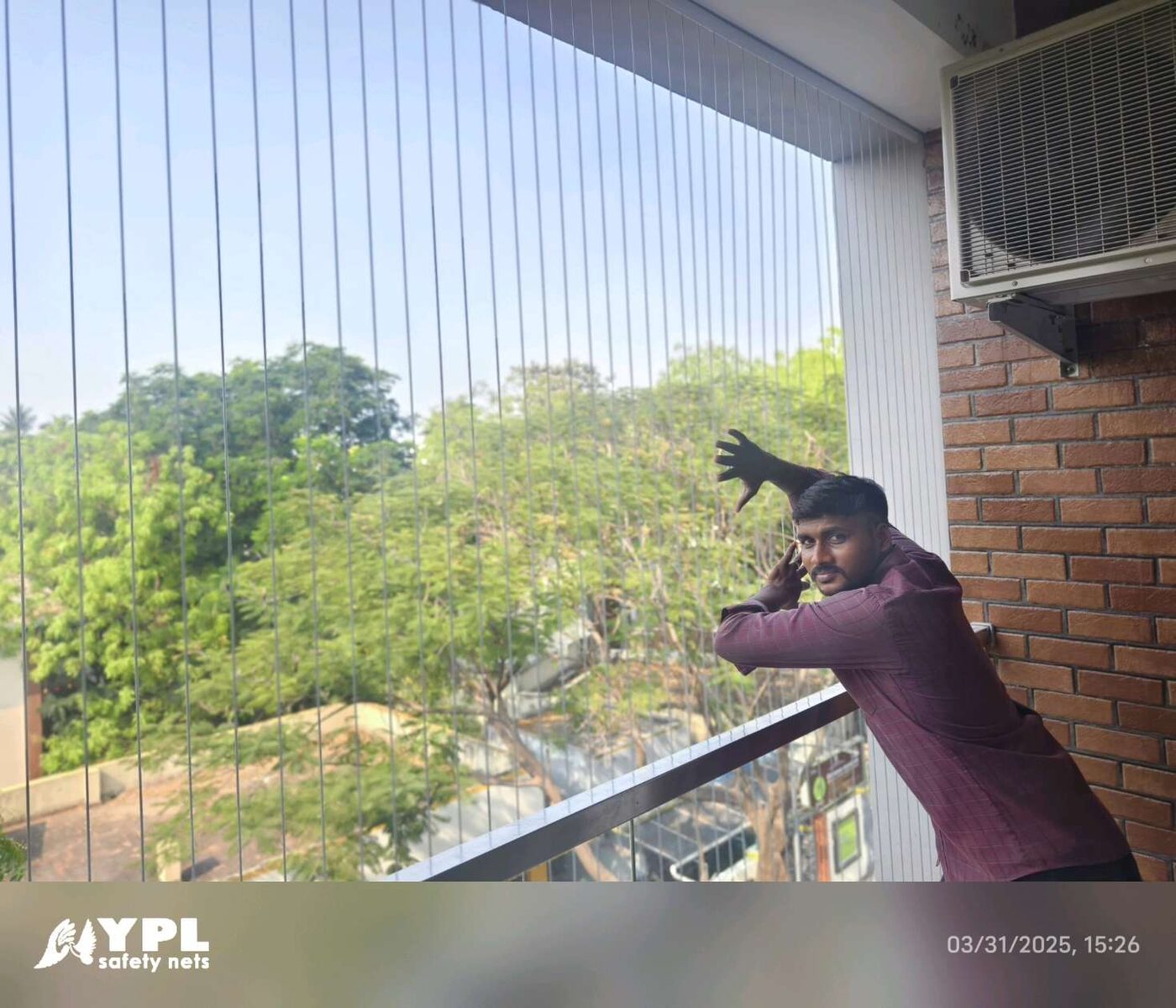 Man beside vertical balcony invisible grills, with trees outside, a brick wall, and an outdoor air conditioner.