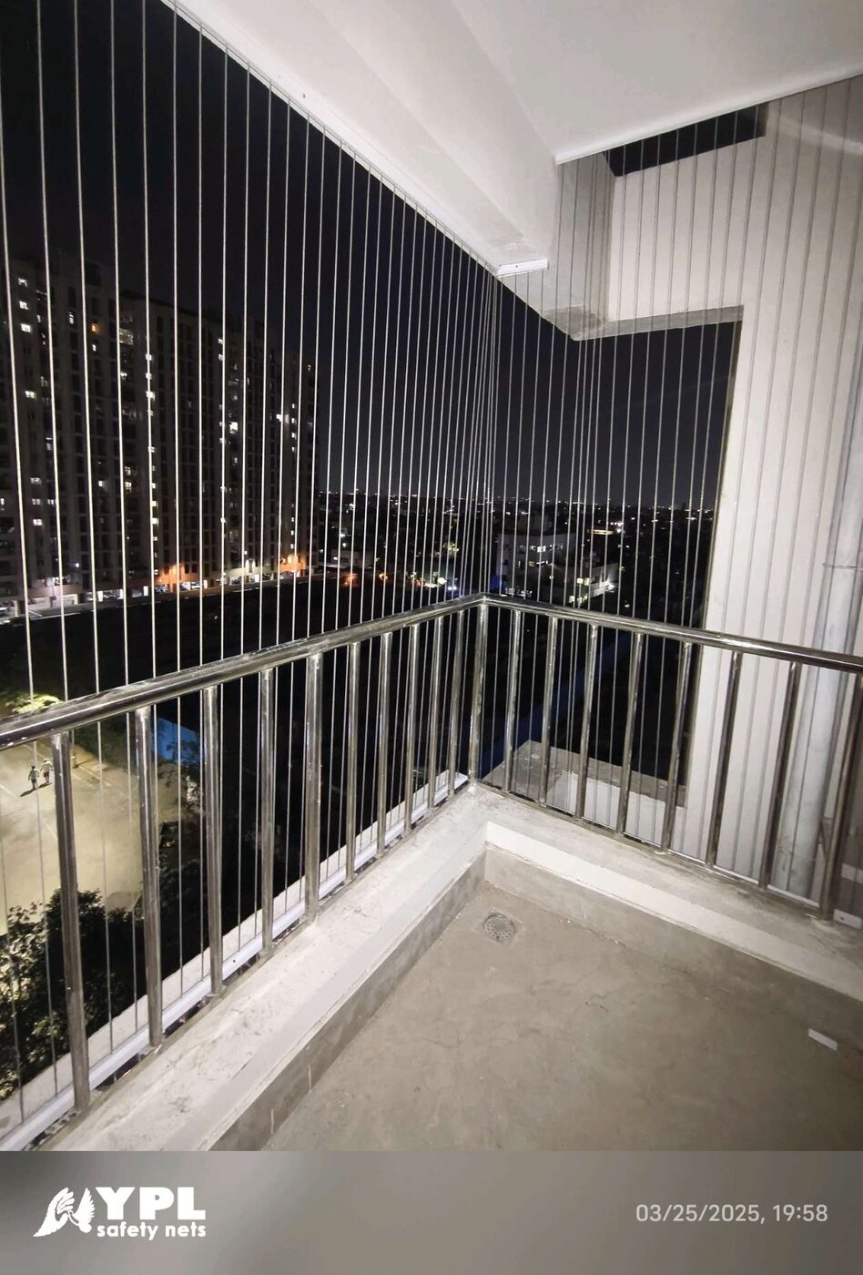 Corner balcony enclosed by vertical invisible grills above metal railings, overlooking neighboring towers and streetlights at night.