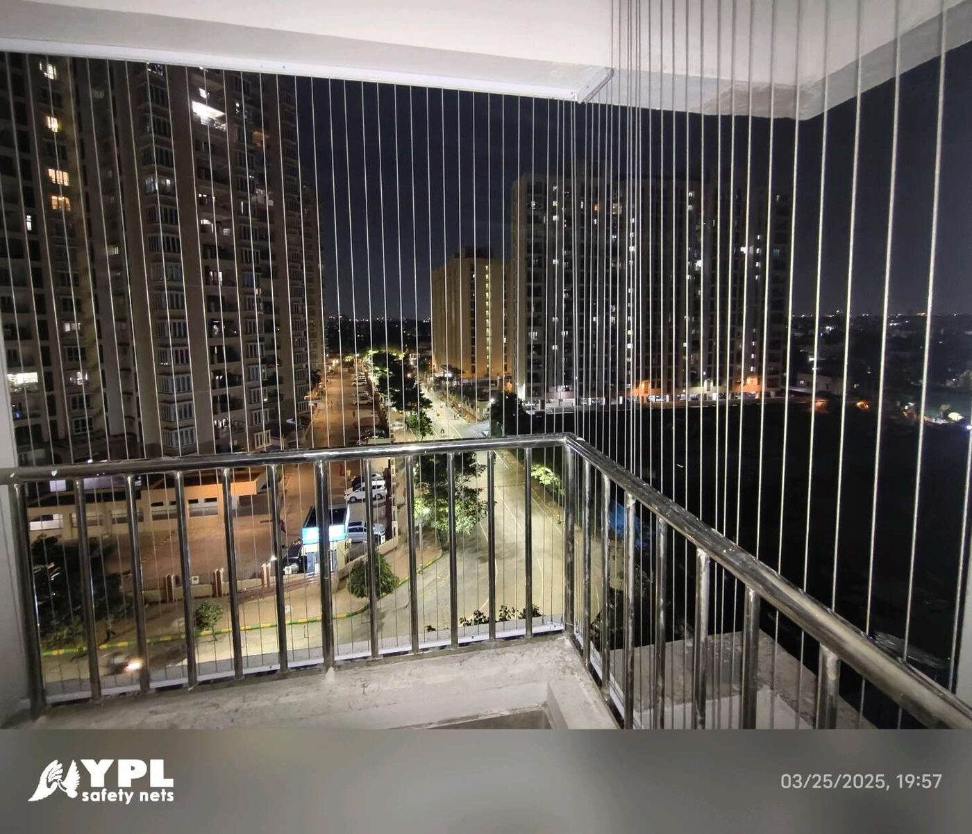 Corner balcony with vertical invisible grills and metal railings overlooking illuminated apartment towers and streets at night.