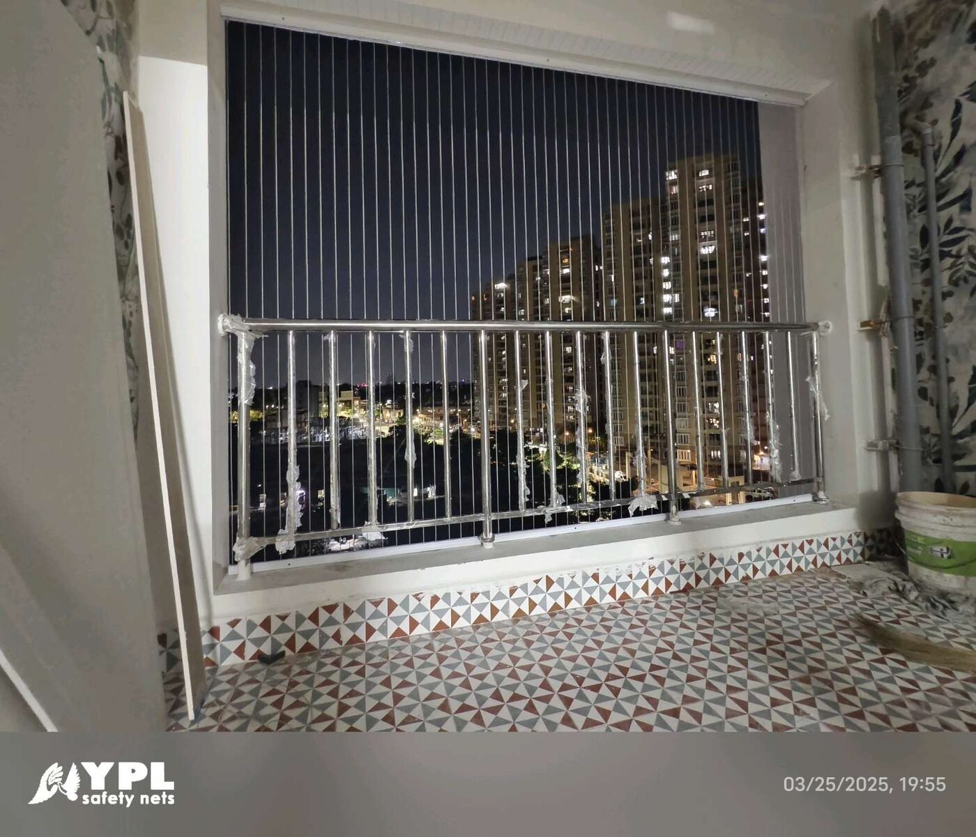 Patterned tile balcony with vertical invisible grills, metal railing, and nighttime views of illuminated apartment towers.