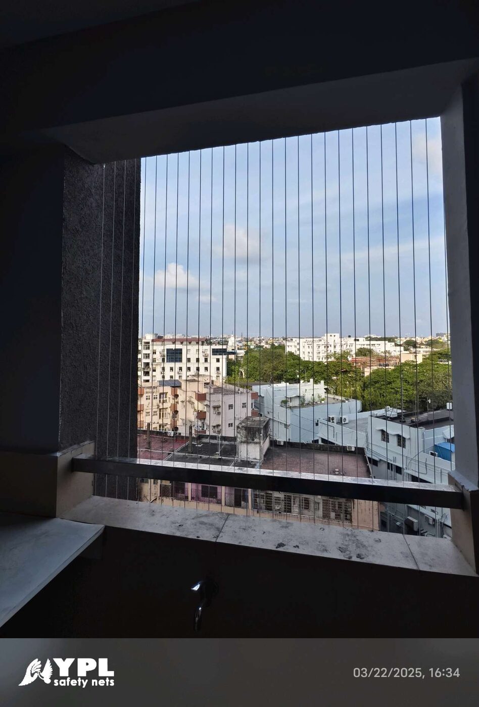 Recessed window opening with vertical invisible grills overlooking neighborhood buildings and trees beneath a blue sky.