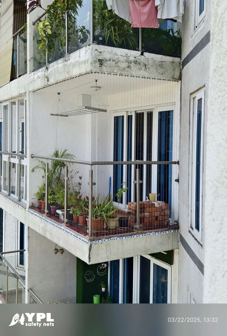 Exterior view of an apartment balcony enclosed with invisible grills, featuring potted plants, hanging drying rack, and stacked bricks.