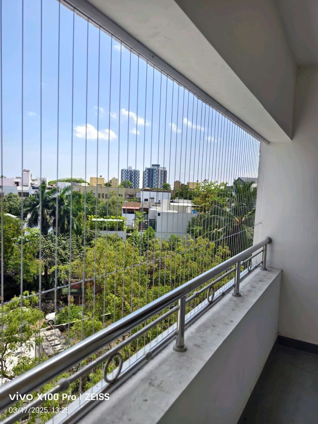 Balcony with vertical invisible grills and stainless steel railing overlooking trees and residential buildings under a bright blue sky.
