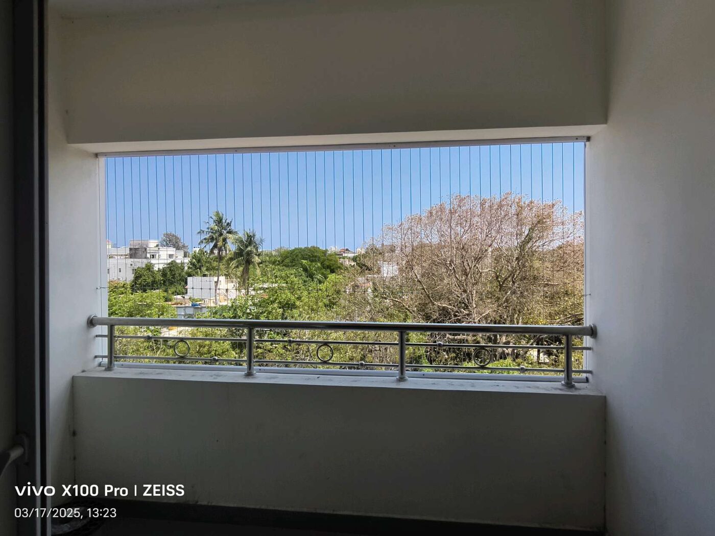 Balcony with vertical invisible grill wires above a metal railing, overlooking trees and nearby buildings.