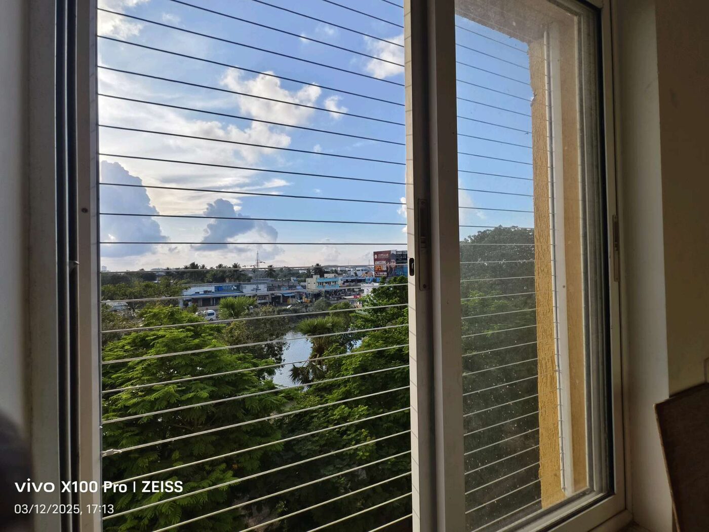 Interior view of a sliding window with horizontal invisible grills overlooking greenery, water, and residential buildings.