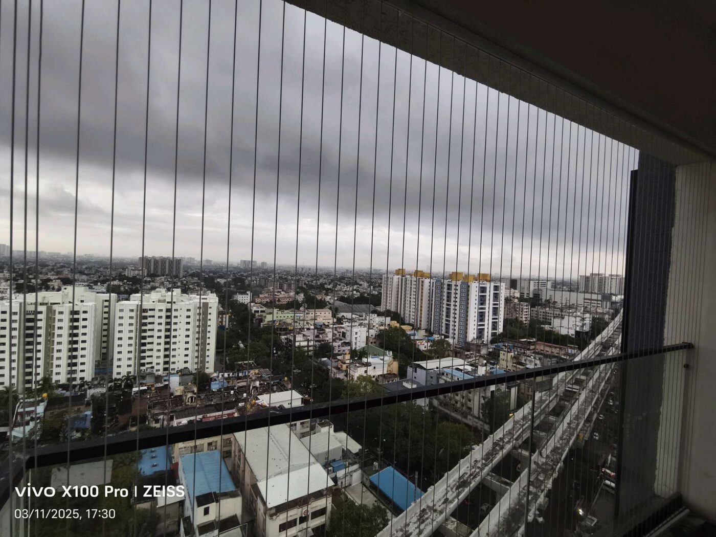 High-rise balcony with vertical invisible grills overlooking apartment blocks and an elevated corridor under dark clouds.