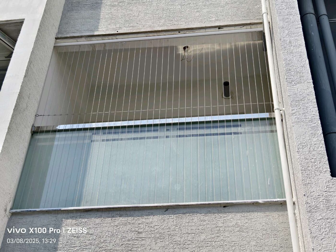 Exterior view of a recessed balcony with vertical invisible grill wires, frosted glass railing, and adjacent pipes.