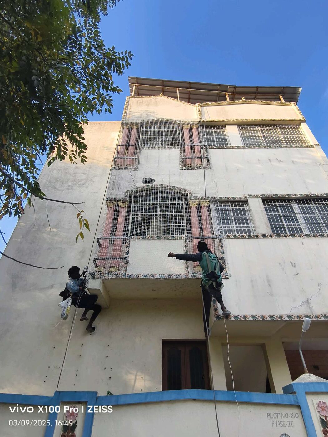 Workers suspended by ropes install protective mesh outside grilled windows on a residential building, viewed from below.