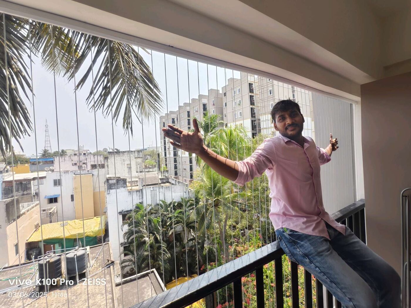 Man seated on a balcony railing behind vertical invisible grills, overlooking palm trees and neighboring buildings.