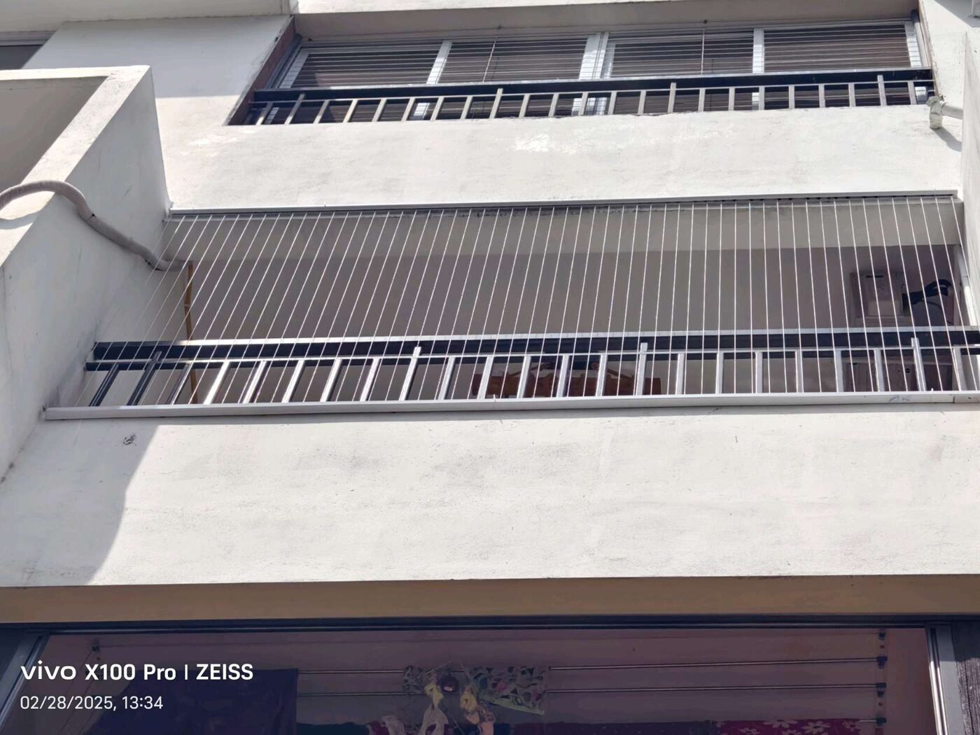 Exterior view looking up at a white building’s balcony fitted with vertical invisible grills above a black railing.