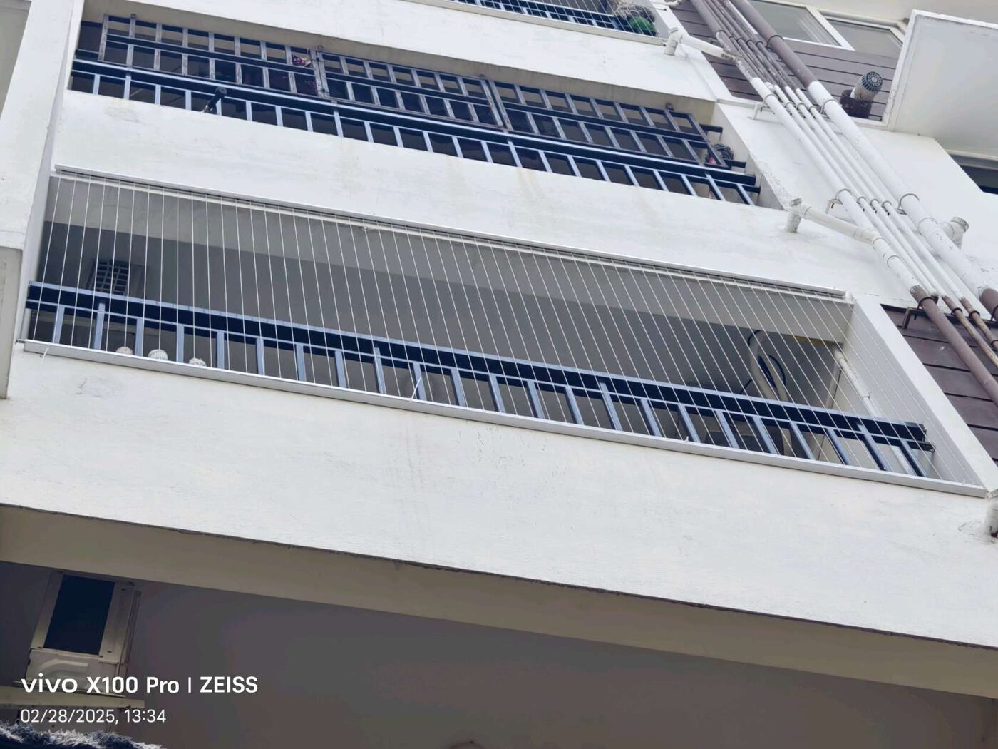 Upward exterior view of stacked apartment balconies, with vertical invisible grills enclosing the lower balcony.