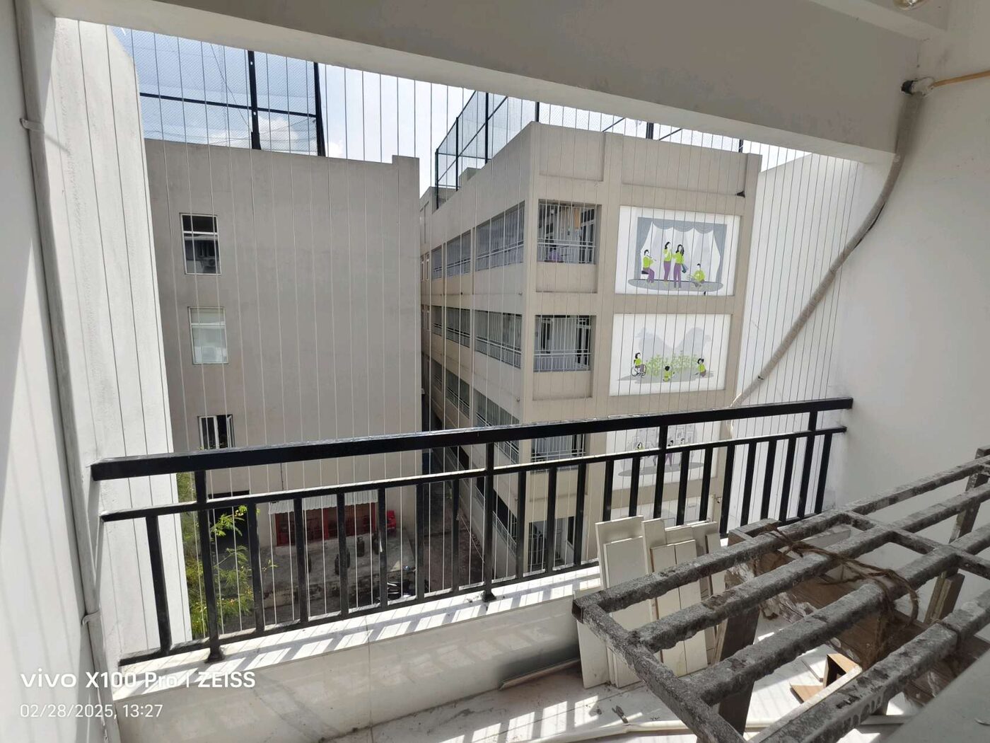 Balcony with vertical invisible grills and black railing facing neighboring buildings, with construction equipment in the foreground.