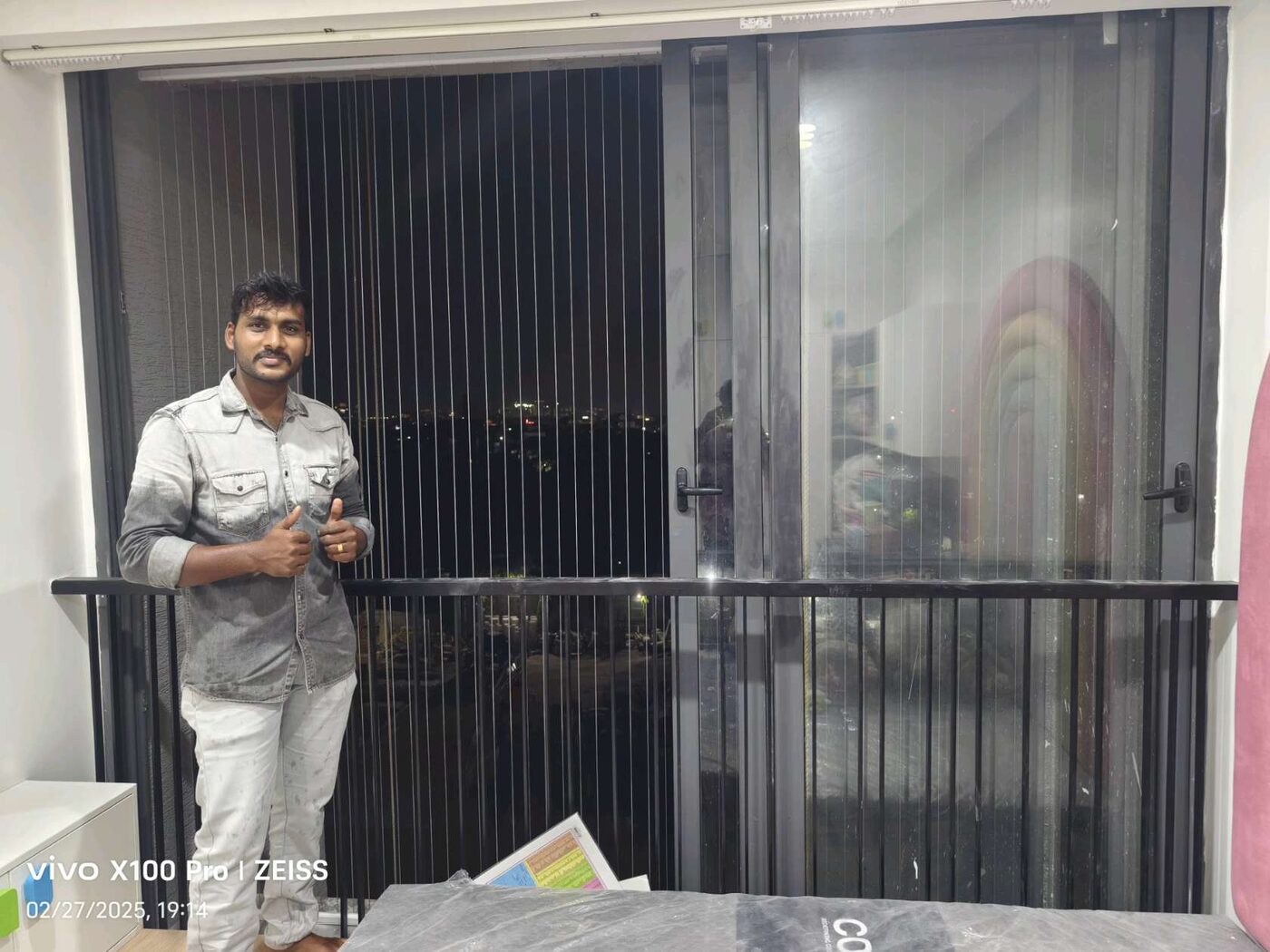 Man standing beside a large sliding window fitted with vertical invisible grills and a black railing at night.