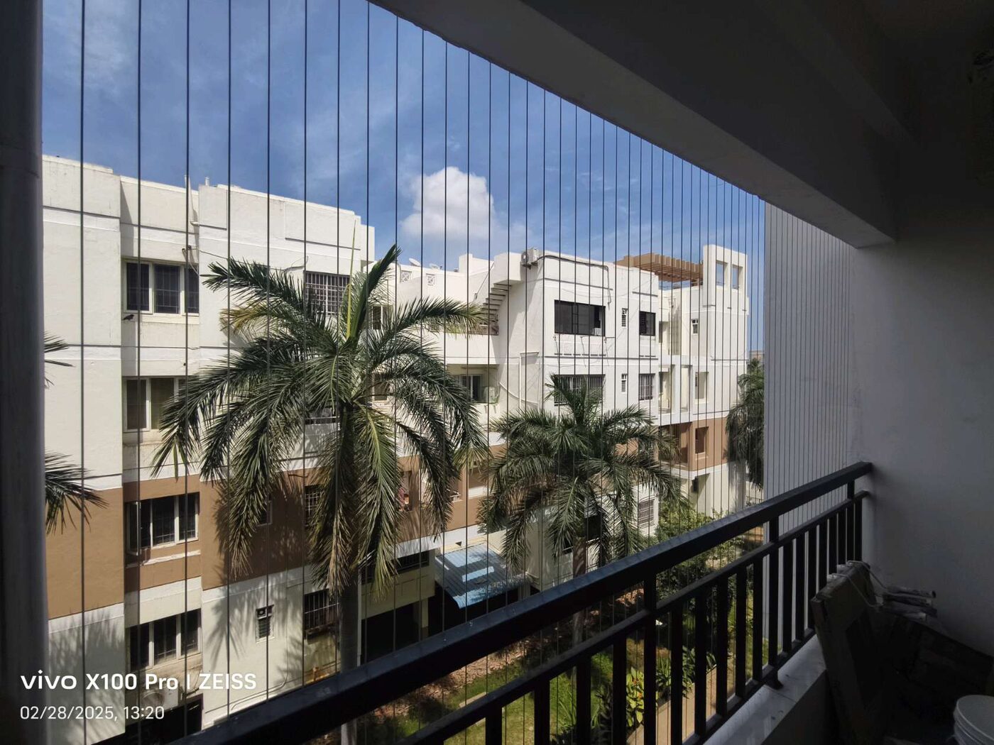 Balcony with vertical invisible grill wires and black railing overlooking palm trees and neighboring apartments.