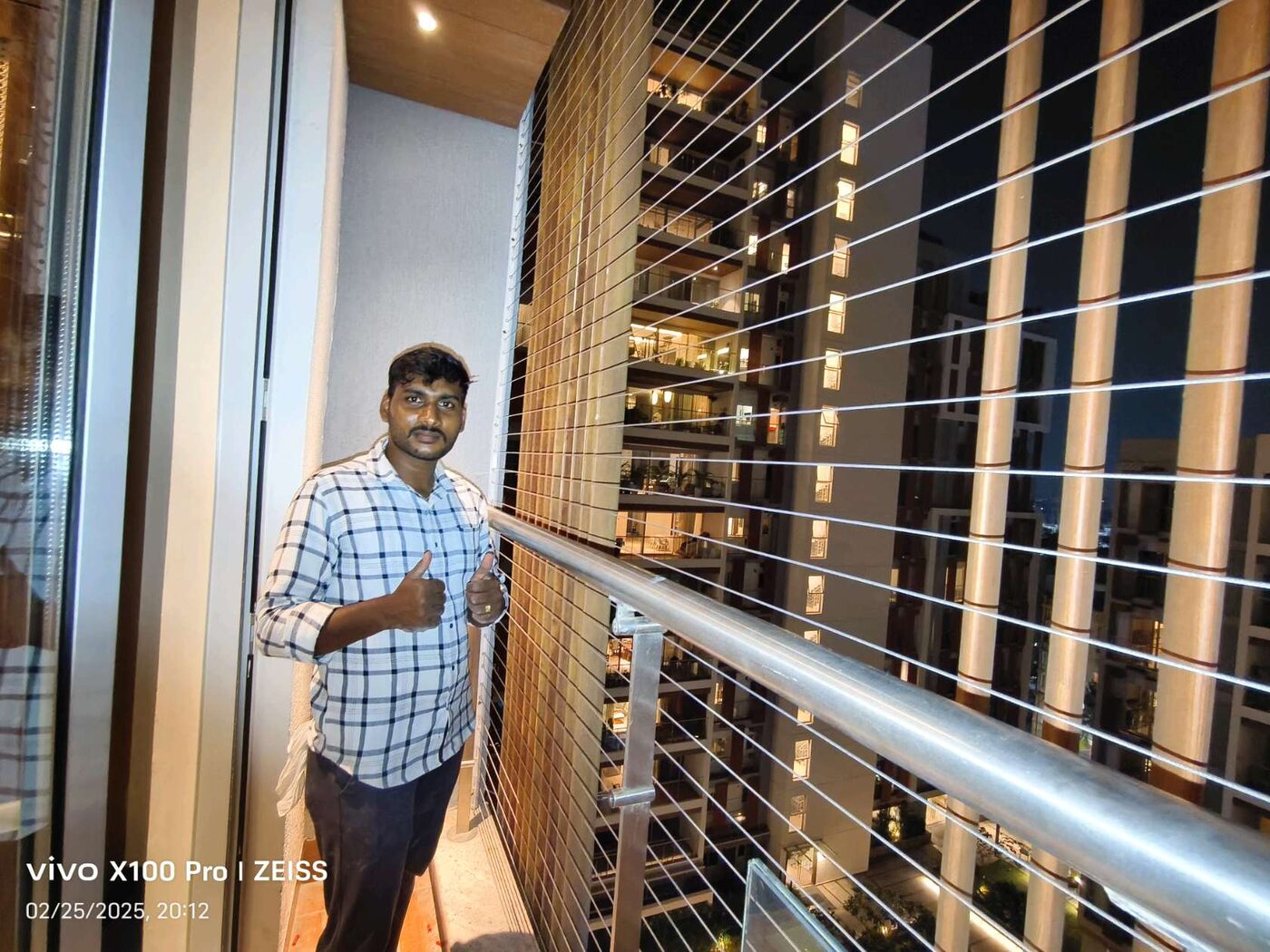 Man giving thumbs-up on a narrow balcony with horizontal invisible grills and illuminated neighboring towers at night.