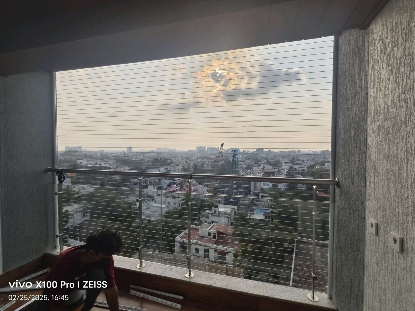 Balcony opening fitted with horizontal invisible grills above a glass railing, overlooking a cityscape beneath cloudy skies.