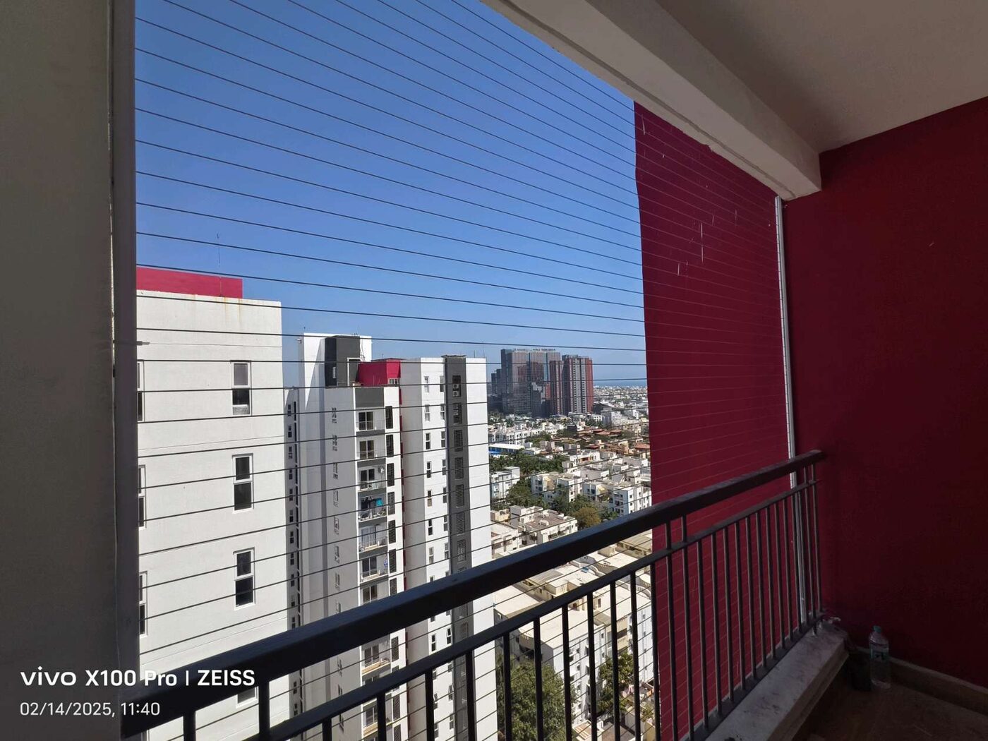 Red-walled balcony with horizontal invisible grills and black railing overlooking apartment towers and a distant coastline.