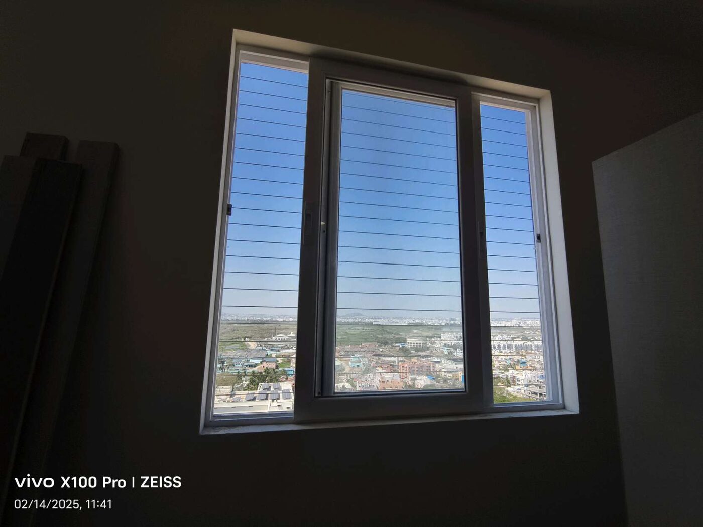 White-framed window with horizontal invisible grills, viewed from a dim interior overlooking buildings and open land.