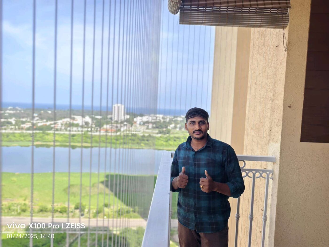 Man giving thumbs up on a balcony enclosed with vertical invisible grills, overlooking a lake and greenery.