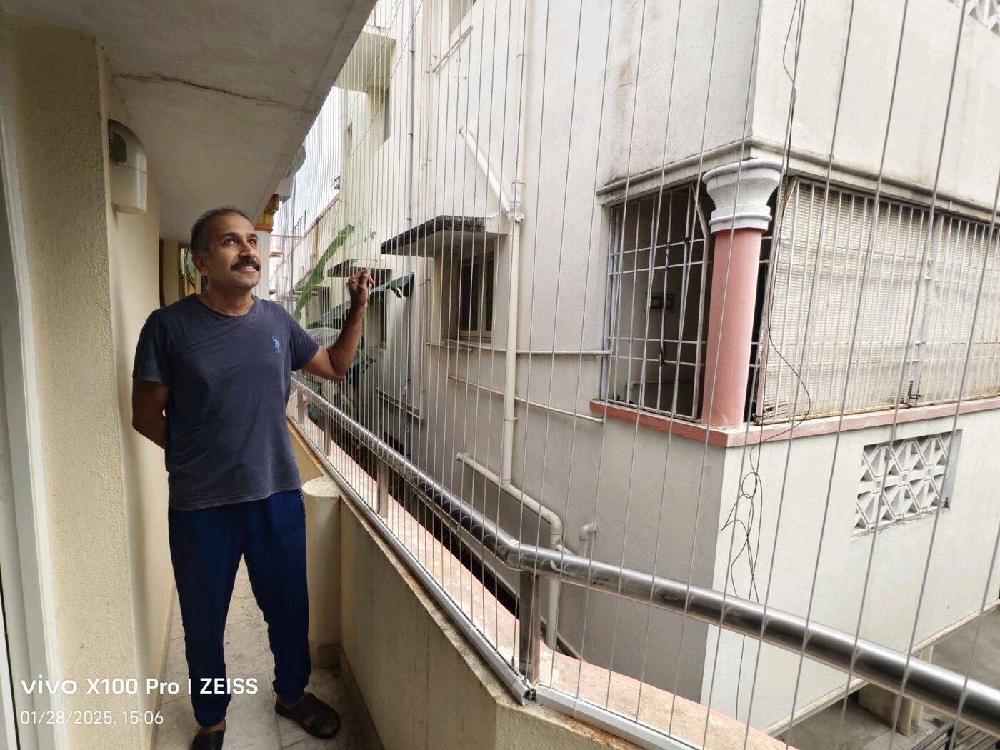 Man standing beside vertical invisible grills on a narrow balcony facing a neighboring building.