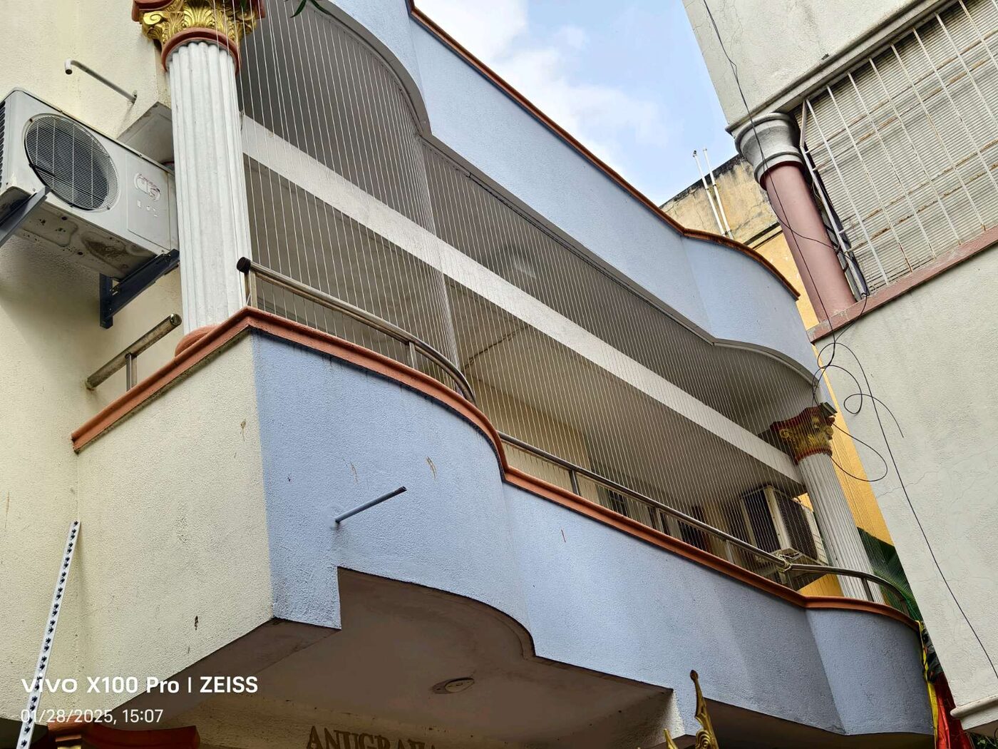 Exterior view of a curved balcony enclosed with vertical invisible grills, flanked by decorative columns and neighboring buildings.