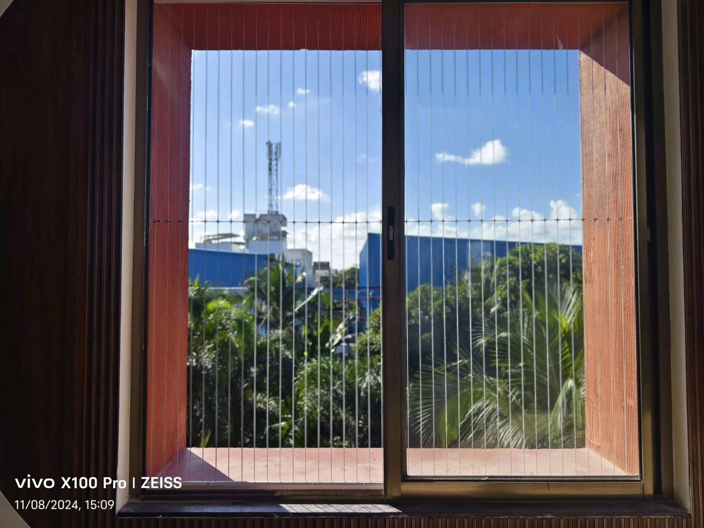 Sliding window with vertical invisible grills, framing trees, a blue building, and cloudy blue sky.